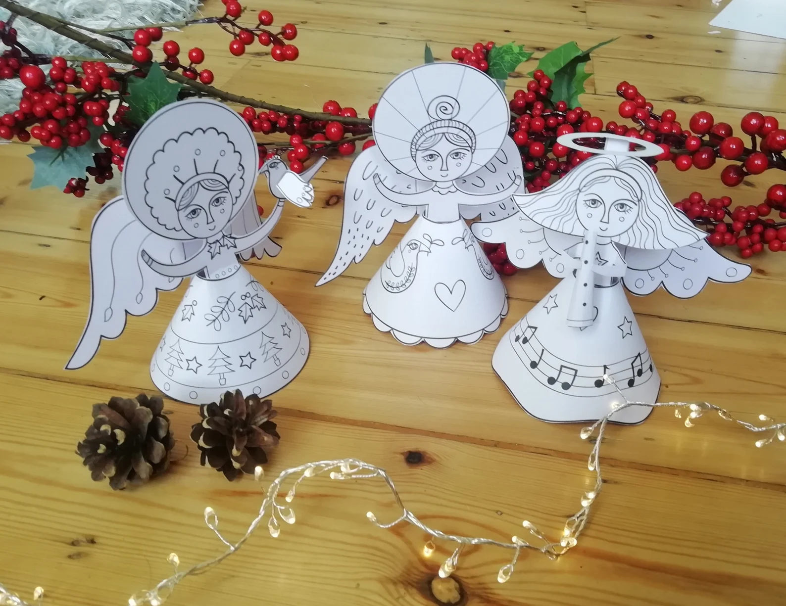 White paper angel cutouts on a wooden surface with red berries and string lights.